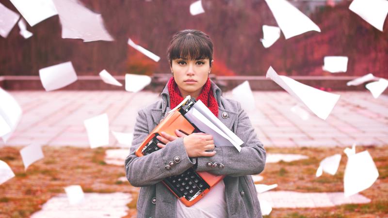 A woman with a cold, dead stare clutching a typewriter to her chest. Blank pieces of paper are falling from the sky around her.